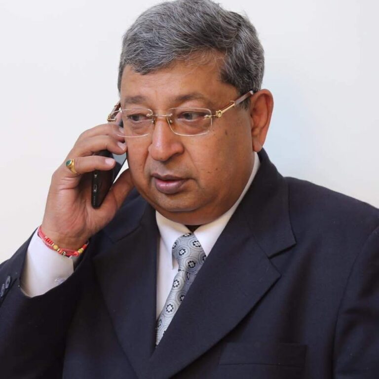 A dignified man in a suit speaking on a mobile phone, wearing glasses and a tie, possibly a school principal or administrator in a formal setting.